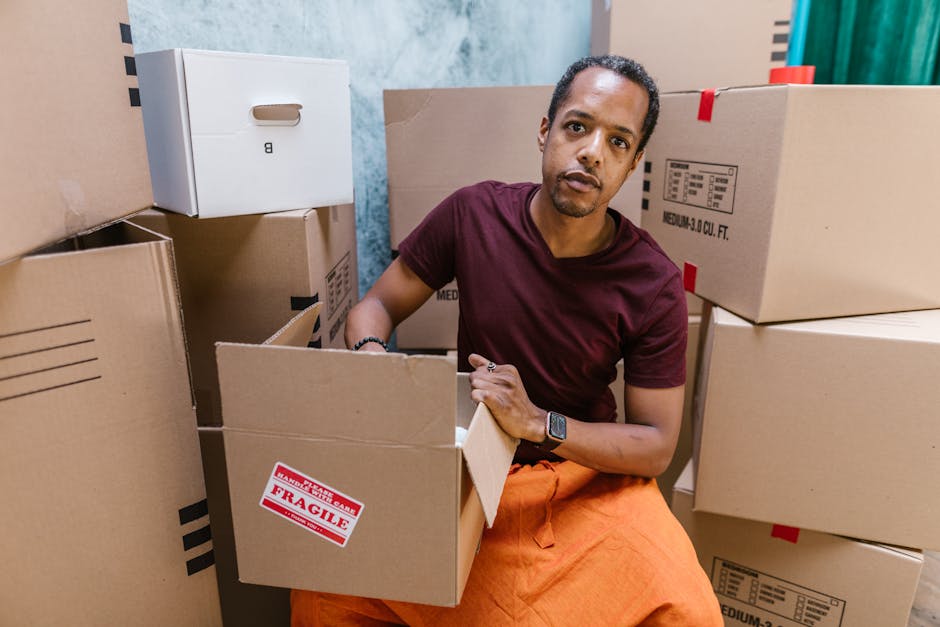 A man with dark curly hair and medium skin tone is seated on the floor among stacked cardboard boxes of various sizes in a room. He is wearing a maroon t-shirt, orange pants, and a black smartwatch on his left wrist. The boxes are made of brown corrugated cardboard, some labeled with red and white 'Fragile' stickers and others with printed specifications. The boxes are arranged on the floor, with some leaning against a wall, and a few are open showing packing materials inside. Behind the man, there are additional sealed boxes and a partial view of a doorway or opening. The environment appears to be during a home relocation or move-in process, with clear focus on furniture transport and packing tasks. The scene is well-lit with natural or artificial light, and the setting echoes themes of house removals and moving logistics, as managed by companies such as Man with Van Hounslow.