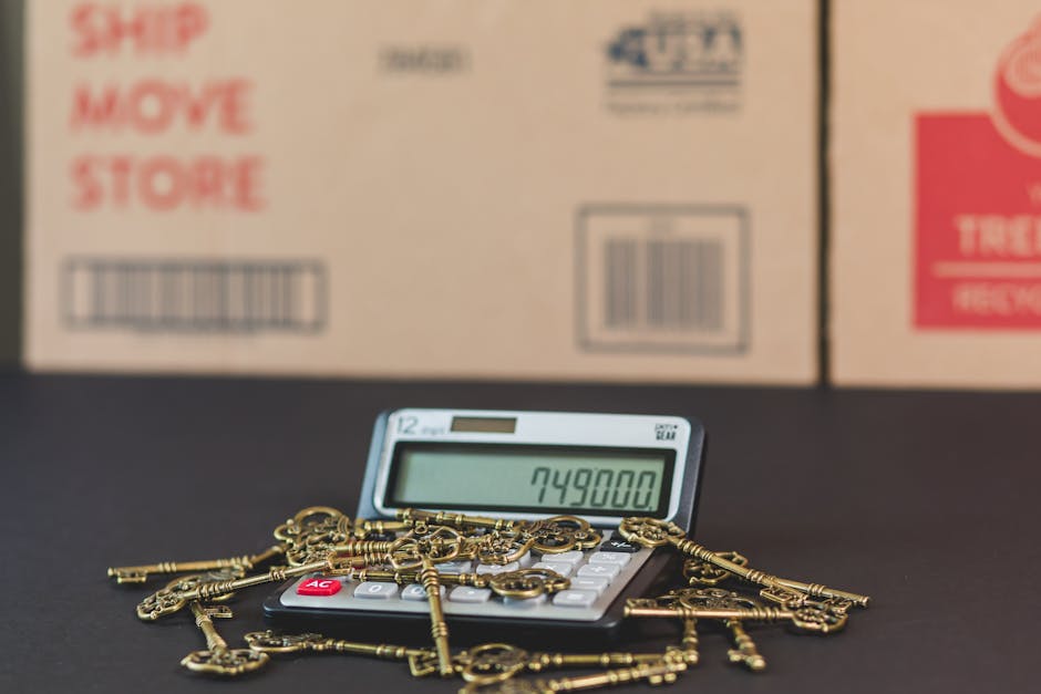 A calculator displaying the number 749800 sits on a black surface, surrounded by several gold-colored keys and keychains. Behind the calculator, there are three large cardboard boxes, partially visible, with printed labels and barcodes. The boxes are situated indoors, in a space likely used for packing or storage during a home relocation process. The environment appears well-lit, and the boxes are arranged in a row, with the middle box being the most prominent. This scene depicts aspects of packing and moving, with an emphasis on careful handling and organization of belongings, typical in furniture transport and house removals. Man with Van Hounslow offers services related to these logistics, ensuring detailed packing and efficient loading, as suggested by the context of the boxes and the packing accessories visible in the image.