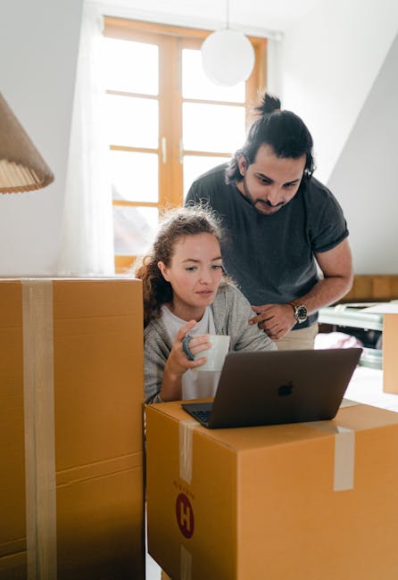 A man with dark hair tied back in a ponytail and a mustache, wearing a dark grey t-shirt, is leaning over a young girl with curly brown hair who is sitting at a table in a well-lit room with large windows and wooden frames. The girl is holding a white mug and looking at a laptop placed on top of a cardboard moving box, which is taped shut with clear packing tape. The scene shows the interior of a house during a home relocation process, with multiple large cardboard boxes stacked nearby, some partially open, indicating ongoing packing and moving activities. The windows behind them let natural light into the room, suggesting daytime. This setting depicts the packing and planning stages of furniture transport and house removals, with clear focus on the activities involved in moving logistics. Man with Van Hounslow is associated with professional removals, supporting home relocation services.