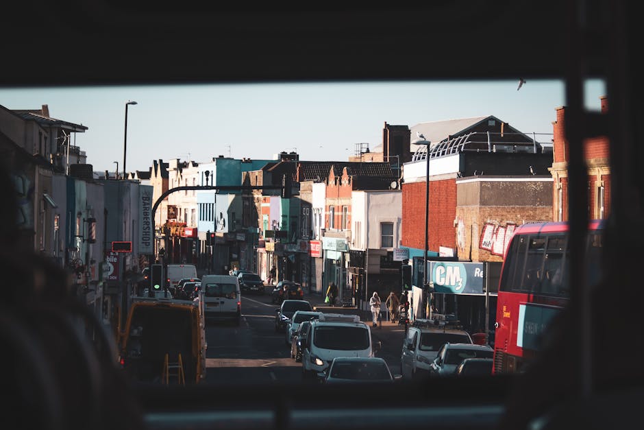 View of a busy urban street captured from inside a vehicle, with the foreground showing the dark interior frame of the bus window. Outside, there are several parked and moving cars on the road, including a white van and a red double-decker bus. Pedestrians walk along the sidewalk lined with small retail shops and businesses, some with colorful storefront signs. The buildings are a mix of brick and painted facades, varying in height and architectural style, typical of a town centre. The scene is illuminated by natural daylight, with an overcast sky casting soft shadows. This image highlights the urban environment and traffic conditions that can be encountered during a home relocation or furniture transport in Hounslow, relevant to the services offered by Man with Van Hounslow in facilitating efficient moving routes and parking preparations for house removals from Feltham to Hounslow.