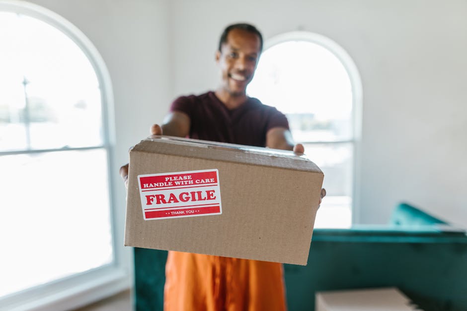 A man with a friendly expression, wearing a maroon t-shirt and orange trousers, is indoors near large arched windows with natural light. He is holding a medium-sized cardboard box with both hands, which has a red and white label that reads 'Please Handle With Care, Fragile.' The setting appears to be a home interior, possibly a living room or hallway, with visible furniture and walls in the background. This image illustrates the careful handling and packing process involved in home relocation or furniture transport, relevant to household removals services provided by Man with Van Hounslow. It captures the initial stage of loading or moving boxes during furniture transport and packing and moving activities typical of house removals near Hounslow East, Hounslow.