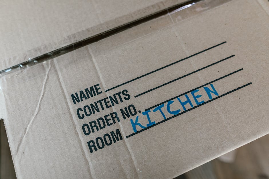 Close-up view of a cardboard moving box with printed labels indicating sections for house relocation, including spaces for the name, contents, order number, and the designated room labeled 'KITCHEN' handwritten in blue ink. The box is sealed with clear packing tape and is positioned inside a property, likely on the floor or countertop, with other packing materials possibly nearby. The background suggests an interior environment, and the image captures the packing process related to a home move managed by Man with Van Hounslow, illustrating the packing and moving preparations associated with house removals and furniture transport services near Hounslow East, Hounslow.