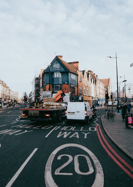 A residential street scene in Hounslow during daytime showing a flatbed truck belonging to Man with Van Hounslow parked on a road with a 20 mph speed limit. The truck is loaded with various furniture items and stacked cardboard boxes, which are secured with straps. A white van is parked behind the truck, and nearby pedestrians and cyclists are present on the pavement. The background features a multi-storey brick building with a blue facade, large windows, and a sloped roof, as well as other commercial and residential buildings lining the street. Streetlights, bicycles, and roadside signage are visible, illustrating typical urban moving logistics for house removals or furniture transport in the area. The scene captures the loading or unloading process involved in home relocation and packing and moving services, supported by the presence of packaging materials and equipment.