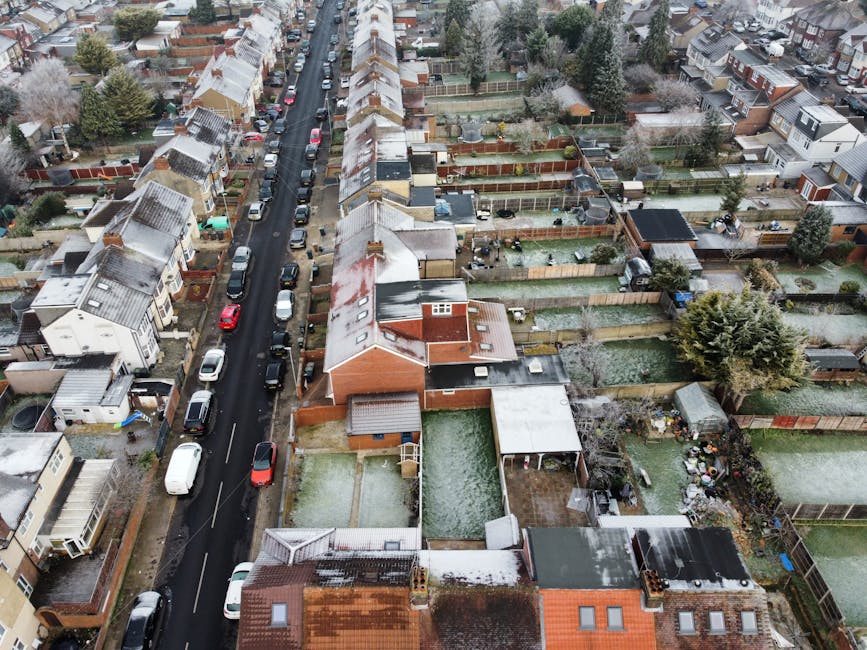 An aerial view of a residential street in Hounslow during winter, showing rows of terraced houses with pitched roofs covered in light snow. Cars are parked along both sides of the narrow asphalt road, which is lined with parked vehicles including a white van. In the foreground, there are back gardens with fences made of wood or metal, some featuring sheds, garden furniture, and small vegetable patches. The houses are a mix of brick and rendered exteriors, with some having extension structures. Tall trees and shrubs are visible throughout the area. This setting illustrates the typical environment for house relocations and furniture transport within urban residential neighborhoods, where careful packing and loading of household items onto vans, such as those operated by Man with Van Hounslow, are common during home relocation or moving services.