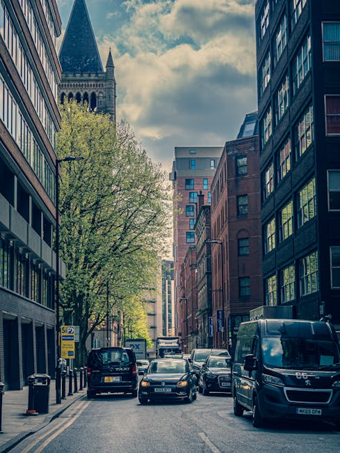 The image shows a narrow urban street scene during daytime with several parked and moving vehicles, including a black van, a black taxi, and a dark-colored car, on both sides of a one-way road in Hounslow. Modern multi-storey office and residential buildings with large windows line the street, with the facades reflecting the partly cloudy sky. A large green tree with fresh spring leaves is situated adjacent to a building on the left side, providing some natural greenery amidst the urban environment. The street features designated parking bays, some occupied by vehicles, and a bus stop with a yellow sign on the left pavement. The scene captures the typical busy feel of a city centre, where local removals or house relocation services, like those offered by Man with Van Hounslow, might be involved in furniture transport or packing and moving activities relying on efficient loading procedures in such a setting.