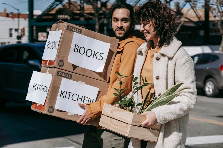 A man and a woman are standing outdoors on a street with parked cars and a brick building in the background, engaged in a home relocation activity. The man, with dark hair and a beard, is holding two cardboard boxes stacked on top of each other, each labeled with white paper indicating contents: the top box labeled 'BOOKS' and the lower box labeled 'KITCHEN' in capital letters. Both boxes are small and sealed with packing tape, designed for item transportation. The woman, with curly brown hair, smiles as she carries a wooden plant box with green leafy plants inside, wrapped with a rubber band. The scene appears to be part of a furniture transport or packing process associated with moving services, with the individuals dressed casually for the task. Bright natural lighting suggests a clear day, and the setting reflects a typical urban environment suitable for a house or flat move managed by [COMPANY_NAME] as part of their removals services, aligning with the TW3 removals guide for Hounslow High Street moves.